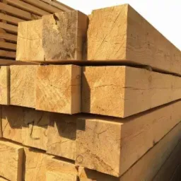 Stos belek z jasnego drewna ułożonych warstwami, widoczne ślady po cięciu, na jednej z belek czerwony znak.