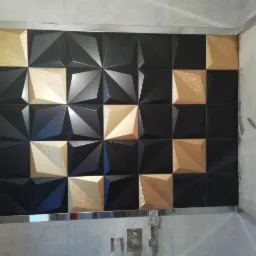 Ściana w łazience z czarno-złotymi, trójwymiarowymi płytkami ułożonymi w szachownicę, wykończona srebrnymi listwami. Widoczny fragment baterii prysznicowej.