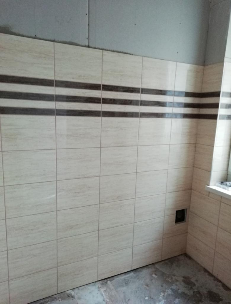 Ściana wyłożona beżowymi płytkami z trzema poziomymi pasami ciemnobrązowych płytek dekoracyjnych; widoczna puszka instalacyjna, surowy sufit i fragment okna.