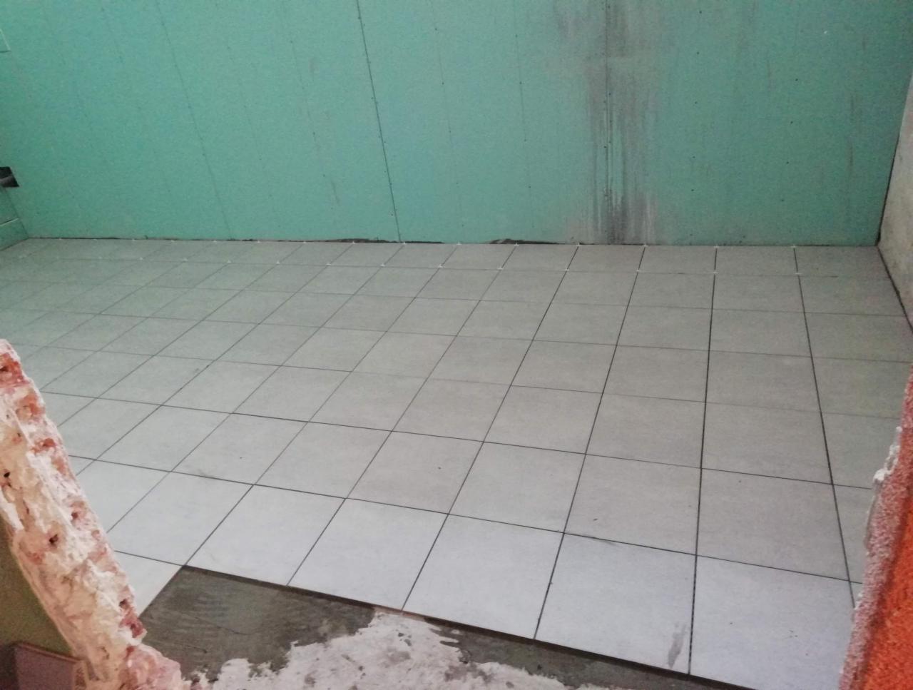 Układanie jasnych, kwadratowych płytek podłogowych w pomieszczeniu z zielonymi ścianami z płyt gipsowo-kartonowych, widoczne resztki zaprawy i gruzu przy krawędziach podłogi.