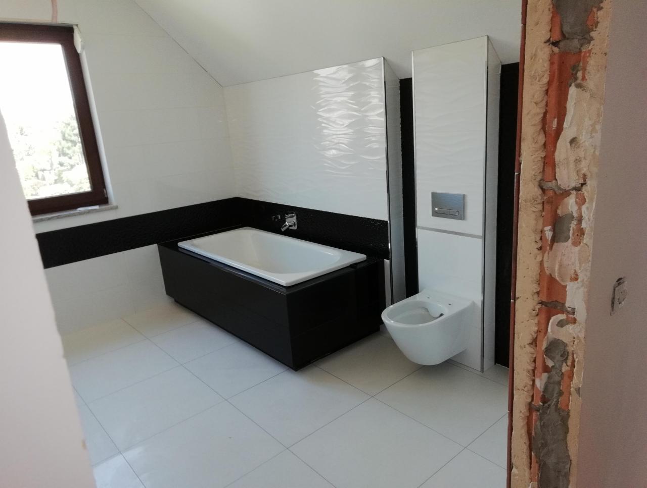 Nowoczesna łazienka w bieli i czerni: wanna z czarną obudową, podwieszana toaleta i białe płytki z delikatnym wzorem. Widoczny fragment surowej ściany w trakcie prac.