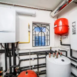 Węzeł cieplny w kotłowni: dwa białe kotły grzewcze, czerwony zbiornik wyrównawczy, liczne czarne rury, manometry i zawory, małe okno z czarną kratą.