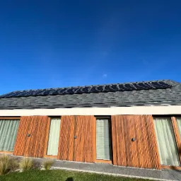 Nowoczesny dom jednorodzinny z panelami słonecznymi na szarym dachu, drewnianą elewacją i dużymi oknami z białymi zasłonami w słoneczny dzień.