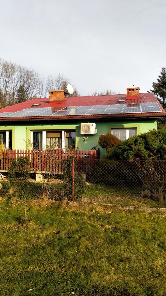 Zielony dom jednorodzinny z czerwonym dachem, na którym zamontowane są panele słoneczne; widoczna antena satelitarna i klimatyzator na ścianie budynku.