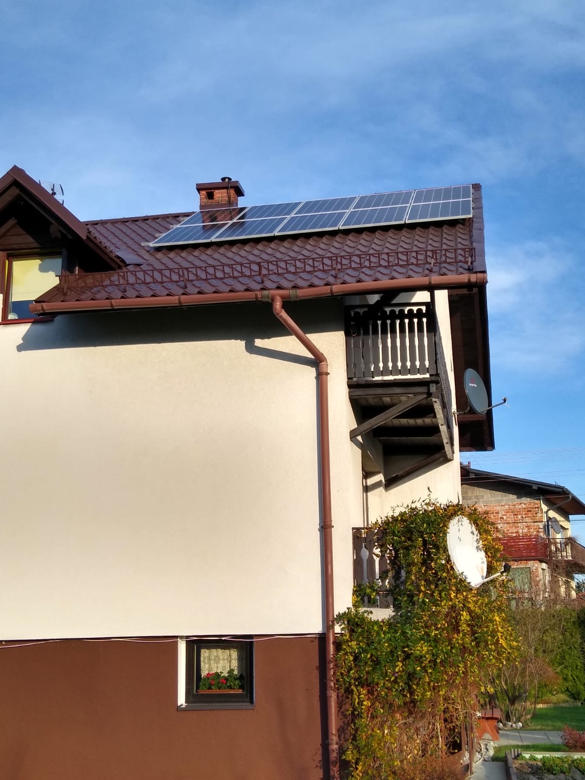 Panele słoneczne na brązowym dachu domu jednorodzinnego z kremową elewacją, widok z dołu w słoneczny dzień.