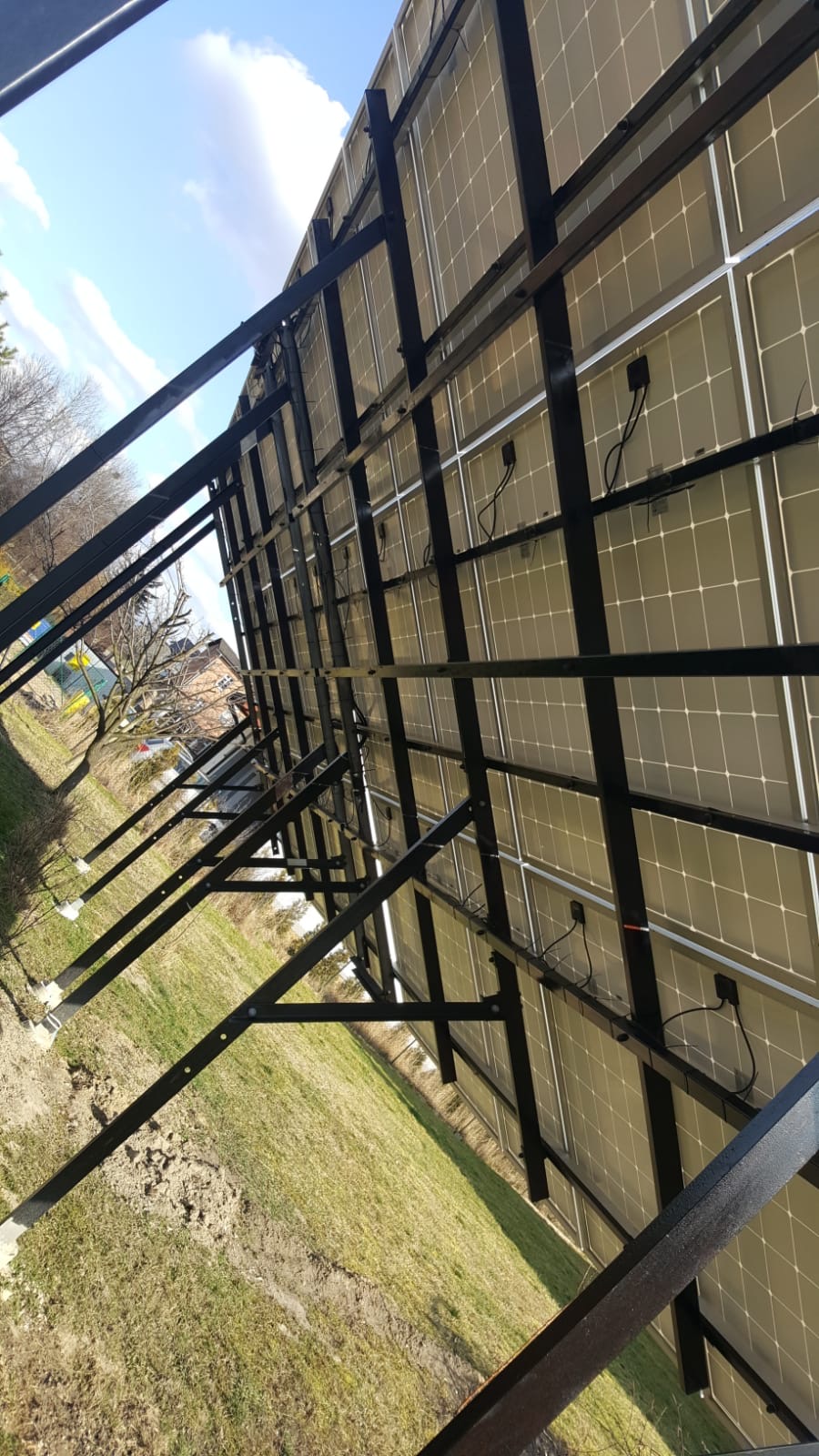 Montaż paneli słonecznych widziany od tyłu, z widoczną konstrukcją wsporczą i okablowaniem, na tle trawnika i błękitnego nieba.