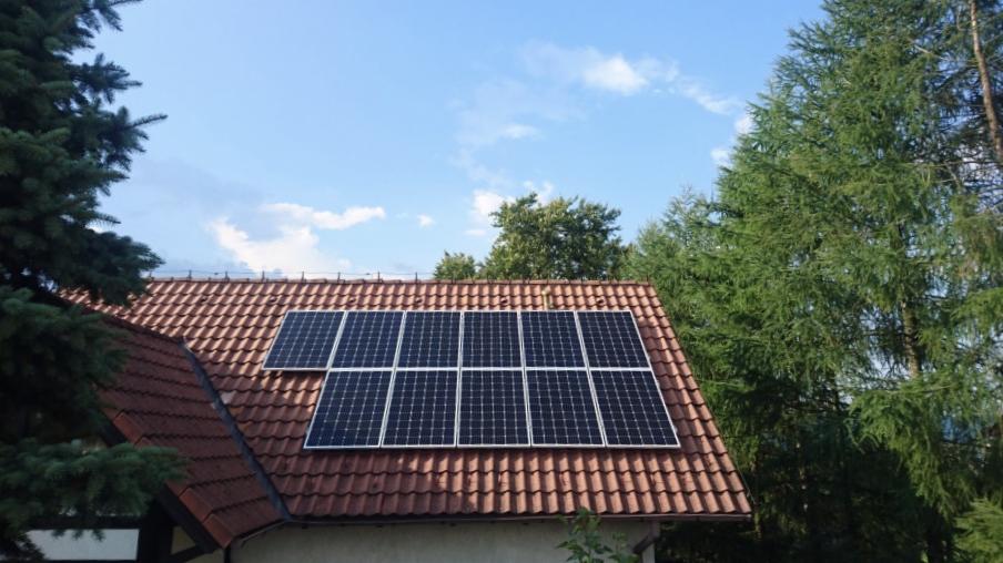 Panel słoneczny na dachu domu z czerwoną dachówką, otoczony zielenią drzew i błękitnym niebem z delikatnymi obłokami.