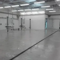 Duża, pusta hala z umytą, mokrą podłogą z szarych płytek, kratka odpływowa na środku, białe kafelki na ścianach, sprzęt przykryty folią ochronną, drabina oparta o ścianę w tle.