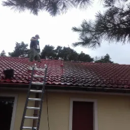 Pracownik na drabinie czyści czerwoną dachówkę domu jednorodzinnego za pomocą myjki ciśnieniowej, widoczna różnica w czystości dachu przed i po czyszczeniu.