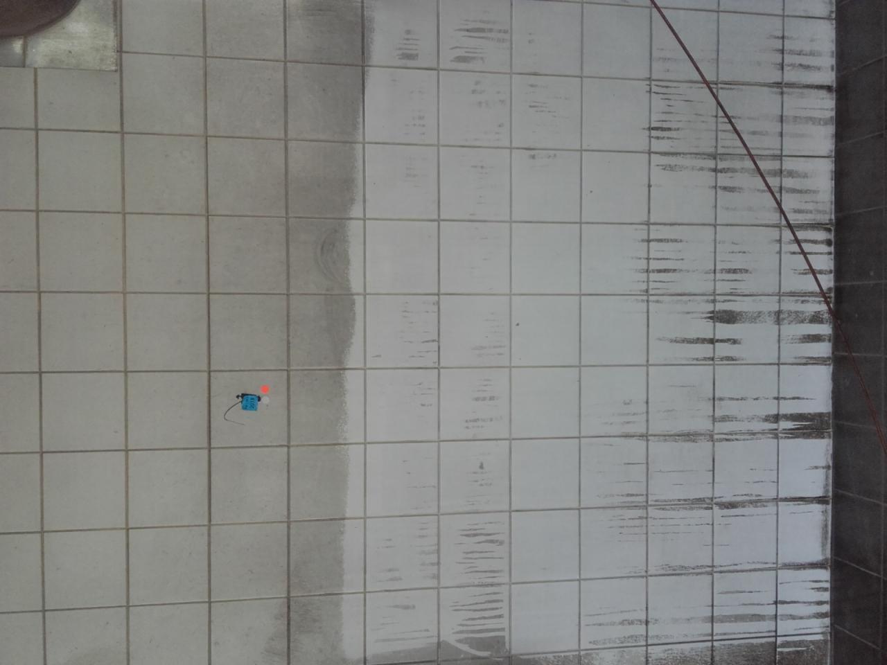 Częściowo umyta podłoga z białych płytek kwadratowych, widoczna różnica w czystości między lewą a prawą stroną, mały niebieski element na jednej z płytek.
