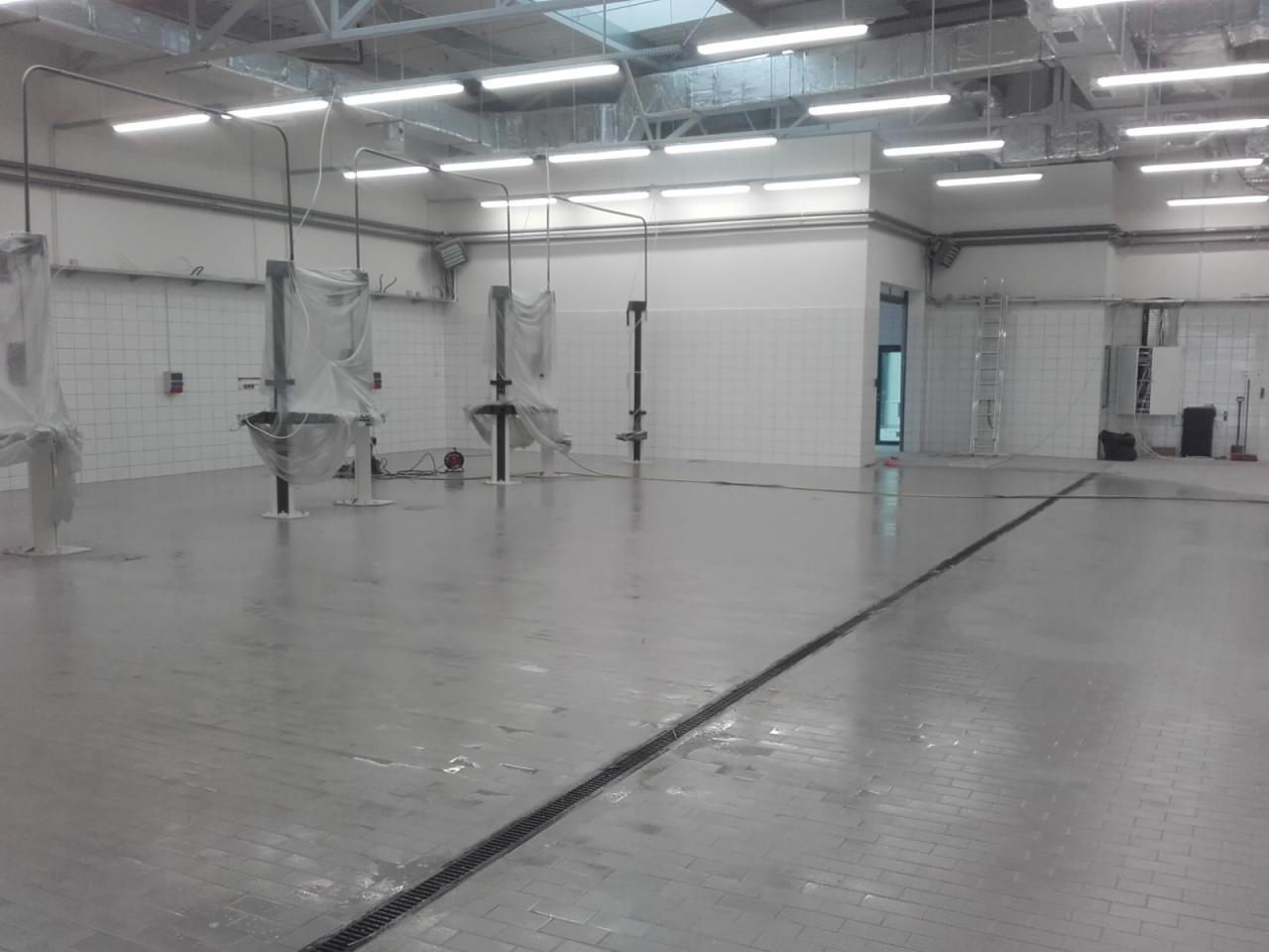 Duża, pusta hala z umytą, mokrą podłogą z szarych płytek, kratka odpływowa na środku, białe kafelki na ścianach, sprzęt przykryty folią ochronną, drabina oparta o ścianę w tle.