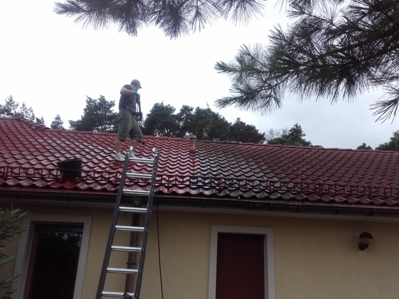 Pracownik na drabinie czyści czerwoną dachówkę domu jednorodzinnego za pomocą myjki ciśnieniowej, widoczna różnica w czystości dachu przed i po czyszczeniu.