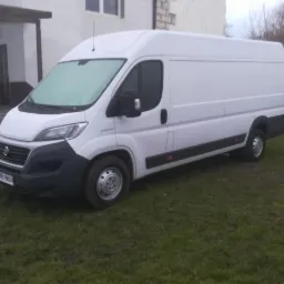 Biały bus dostawczy na trawniku przed budynkiem. Szyba przednia lekko oszroniona. Widoczna tablica rejestracyjna GMB 8W64.