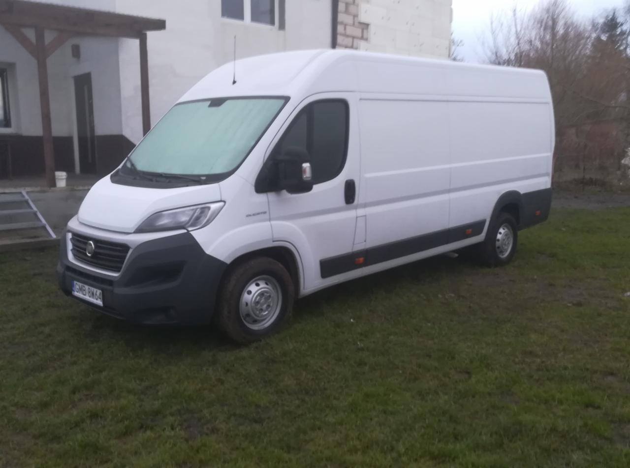 Biały bus dostawczy na trawniku przed budynkiem. Szyba przednia lekko oszroniona. Widoczna tablica rejestracyjna GMB 8W64.