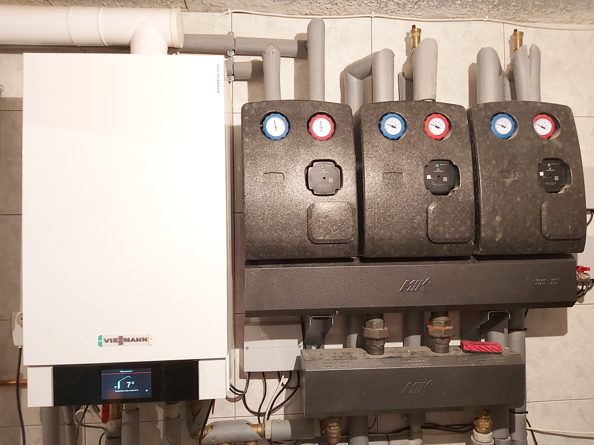Biały gazowy kocioł Vitodens z trzema obiegami grzewczymi, każdy z manometrami i czarnymi obudowami, zamontowany na kafelkowanej ścianie.
