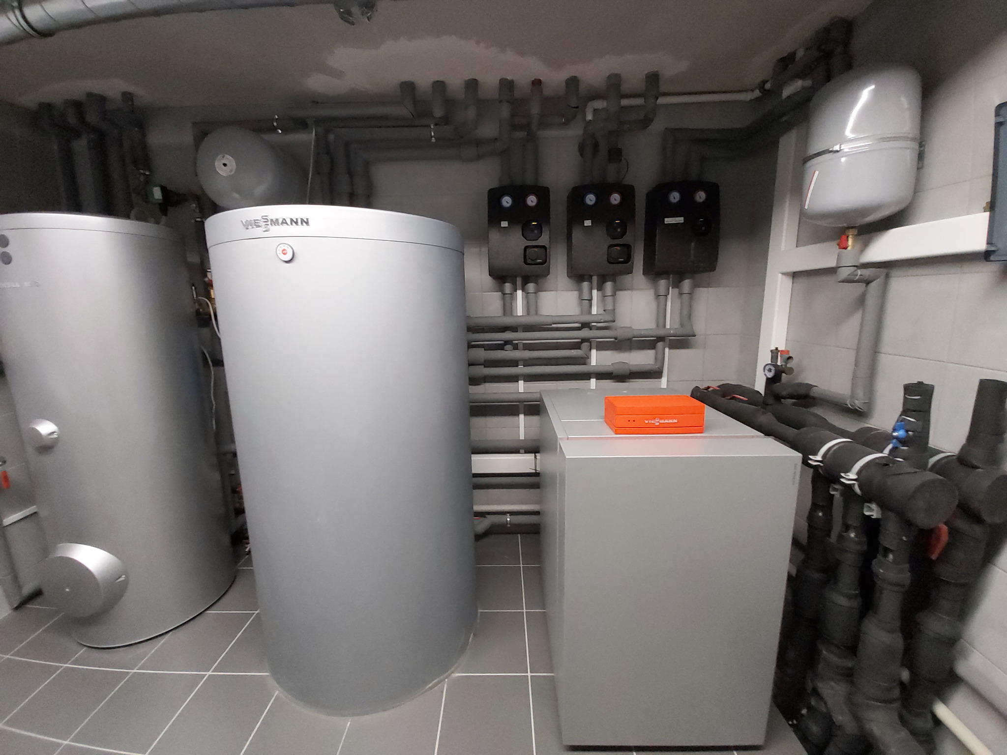 Kompletna instalacja kotłowni z dwoma srebrnymi zasobnikami Viessmann, buforem z pomarańczowym sterownikiem i rozbudowaną siecią rur w szarej kotłowni wyłożonej płytkami.