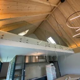 Wnętrze domu z drewnianym stropem i antresolą zabezpieczoną szklaną balustradą mocowaną na metalowych uchwytach, widok na okna dachowe i wiszące lampy w kształcie okręgów, poniżej antresoli...