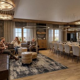 STUDIO INDYWIDUALNYCH INSPIRACJI ARTbis DESIGN Projekty Wnętrz i Elewacji Joanna Wal - Przestronny salon z drewnianą podłogą, skórzaną sofą i długim stołem. Oświetlenie stanowią okrągłe lampy. W tle widoczna kuchnia i siedzące osoby.