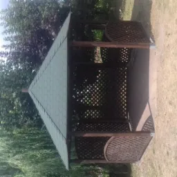 Drewniana altana z ażurowymi ścianami i zielonym dachem na tle drzew i trawnika, widziana z boku.