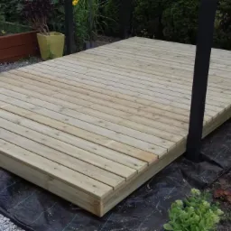 Nowo wybudowany taras z jasnego drewna, wsparty na czarnych słupach, z widocznym podłożem z czarnej agrowłókniny i otaczającą zielenią ogrodu.