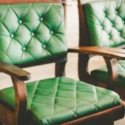 Dwa drewniane fotele z ciemnozieloną, pikowaną tapicerką, widoczne pod kątem, z delikatnym oświetleniem.