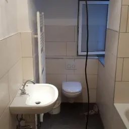 Wnętrze wyremontowanej łazienki w Opolu z umywalką, toaletą i grzejnikiem drabinkowym, widoczne świeże białe płytki na ścianach i ciemna podłoga, kabel od lampy zwisa z sufitu.