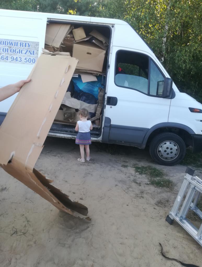 Biały van załadowany kartonami i workami, mała dziewczynka stoi przed otwartymi drzwiami, a ręka trzyma duży arkusz kartonu. W tle drzewa i drabina.