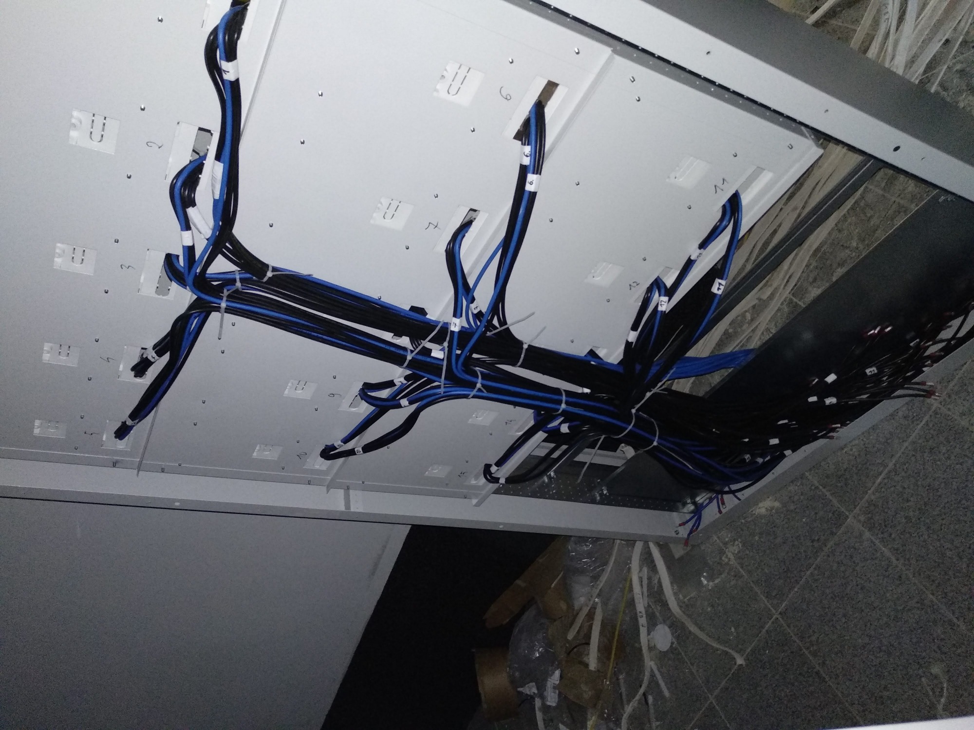 Widok z dołu na podwieszany sufit z zamontowanymi przewodami elektrycznymi, oznaczonymi numerami, częściowo ułożonymi w korytkach kablowych, w trakcie prac instalacyjnych.