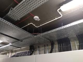 Sufit techniczny z perforowanymi korytami kablowymi, rurami PCV i przewodami, widoczna czujka dymu