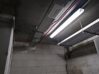 Szara, betonowa ściana z widocznymi blokami, z zamontowanymi metalowymi korytkami kablowymi pod sufitem, oświetlona jarzeniówką. Widoczne czerwone przewody elektryczne.