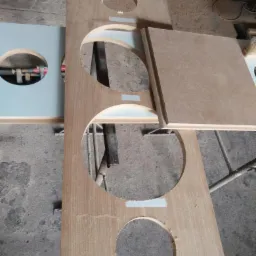 Przygotowywany blat drewniany z wyciętymi otworami, zabezpieczony taśmą malarską, leżący na warsztatowym stole.