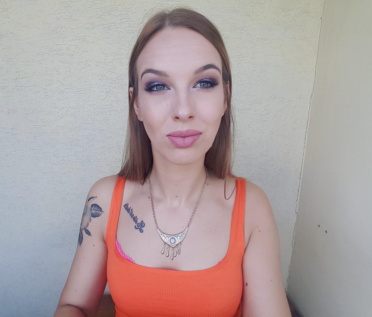 Portret kobiety z makijażem oczu w odcieniach fioletu i różu, w pomarańczowym topie na tle jasnej ściany. Widoczne tatuaże na ramionach i srebrny naszyjnik.