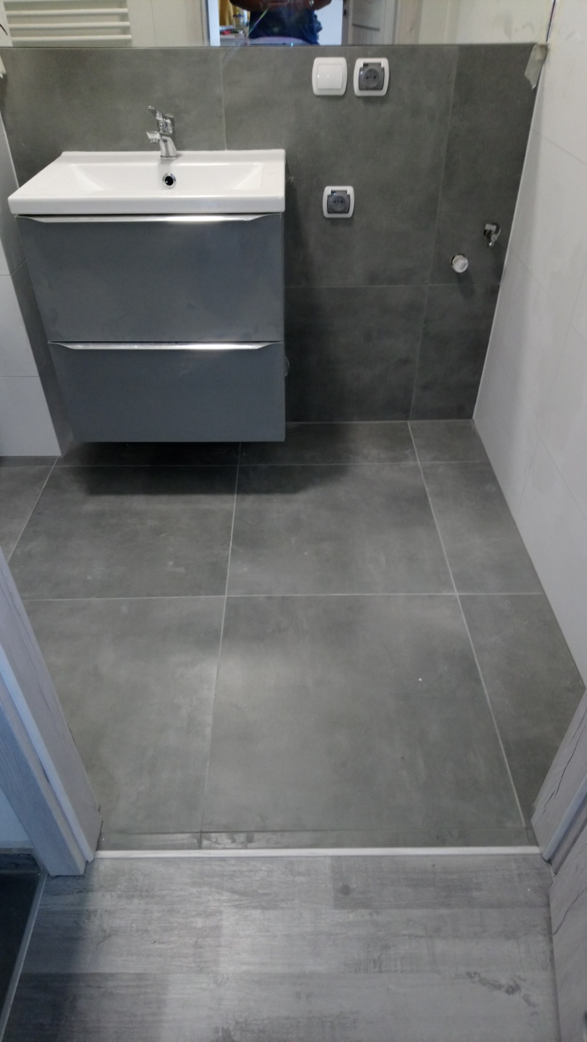 Szare, kwadratowe płytki podłogowe i ścienne w łazience, z umywalką z szafką w kolorze szarym i białym blatem oraz białymi włącznikami i gniazdkami na ścianie.