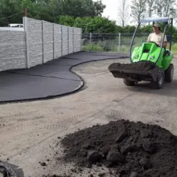 Operator na zielonej miniładowarce rozrzuca ziemię na przygotowanym terenie z czarną geowłókniną i betonowym ogrodzeniem w tle.