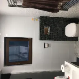 Nowoczesna łazienka z białymi płytkami, mozaiką z czarnych płytek za toaletą, oknem z brązową ramą i grzejnikiem drabinkowym z brązowym ręcznikiem.