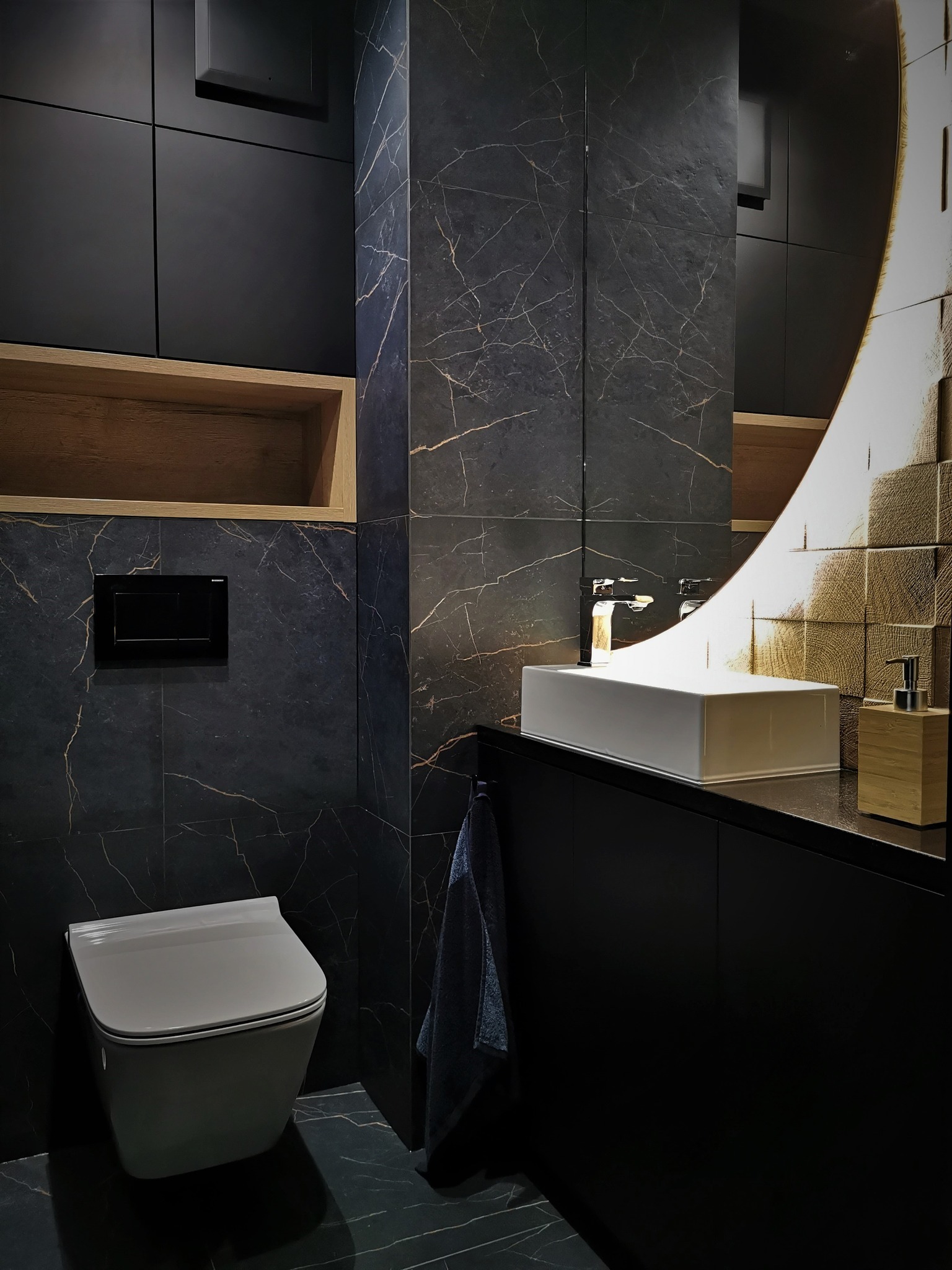 Nowoczesna łazienka w ciemnej tonacji z marmurowymi płytkami, podświetlanym lustrem i wiszącą toaletą. Minimalistyczny design z drewnianymi akcentami.