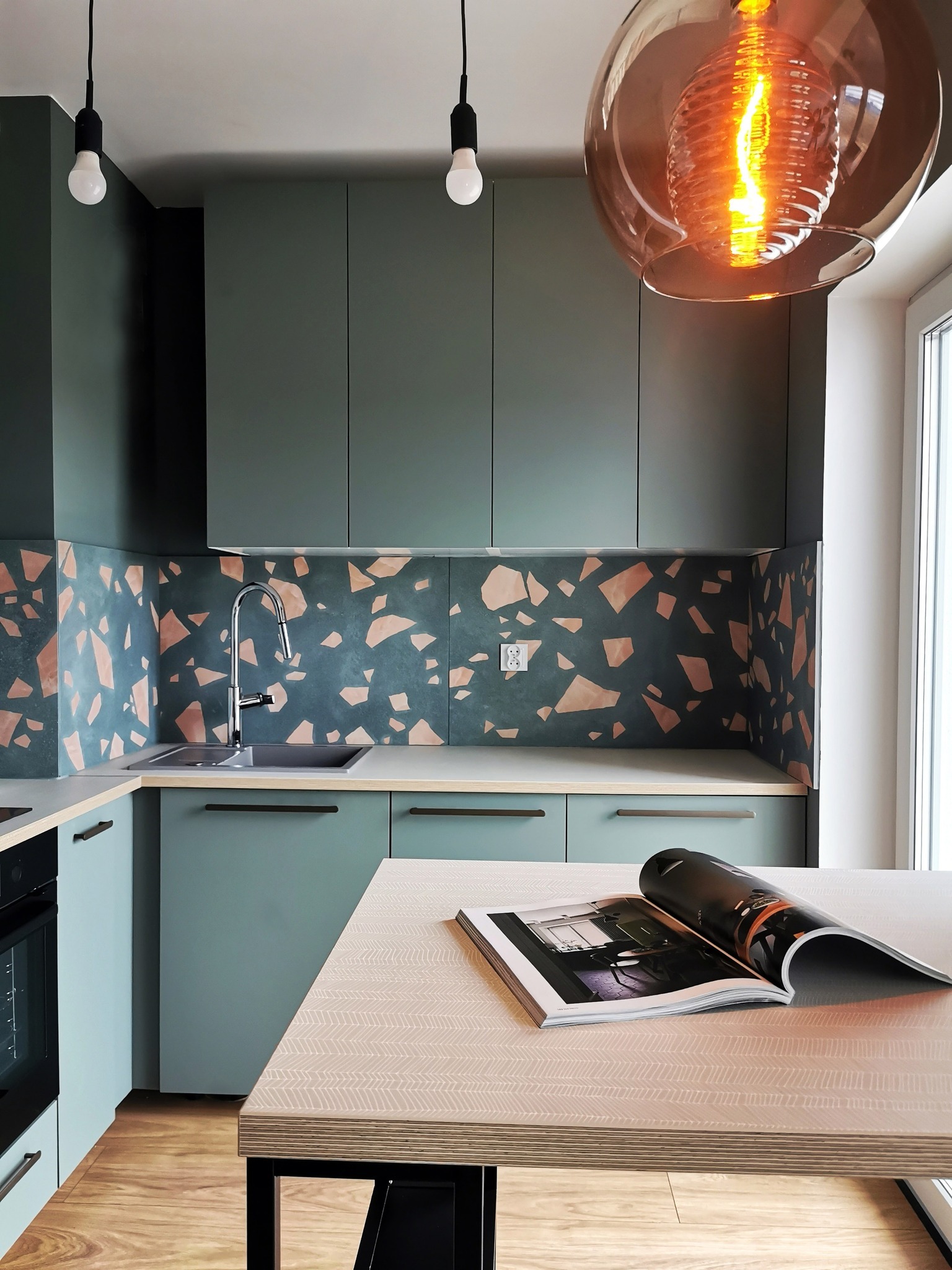 Nowoczesna kuchnia w odcieniach zieleni i terazzo. Uwagę przykuwa designerska lampa wisząca i otwarty magazyn na blacie. Minimalistyczne fronty szafek i drewniana podłoga.