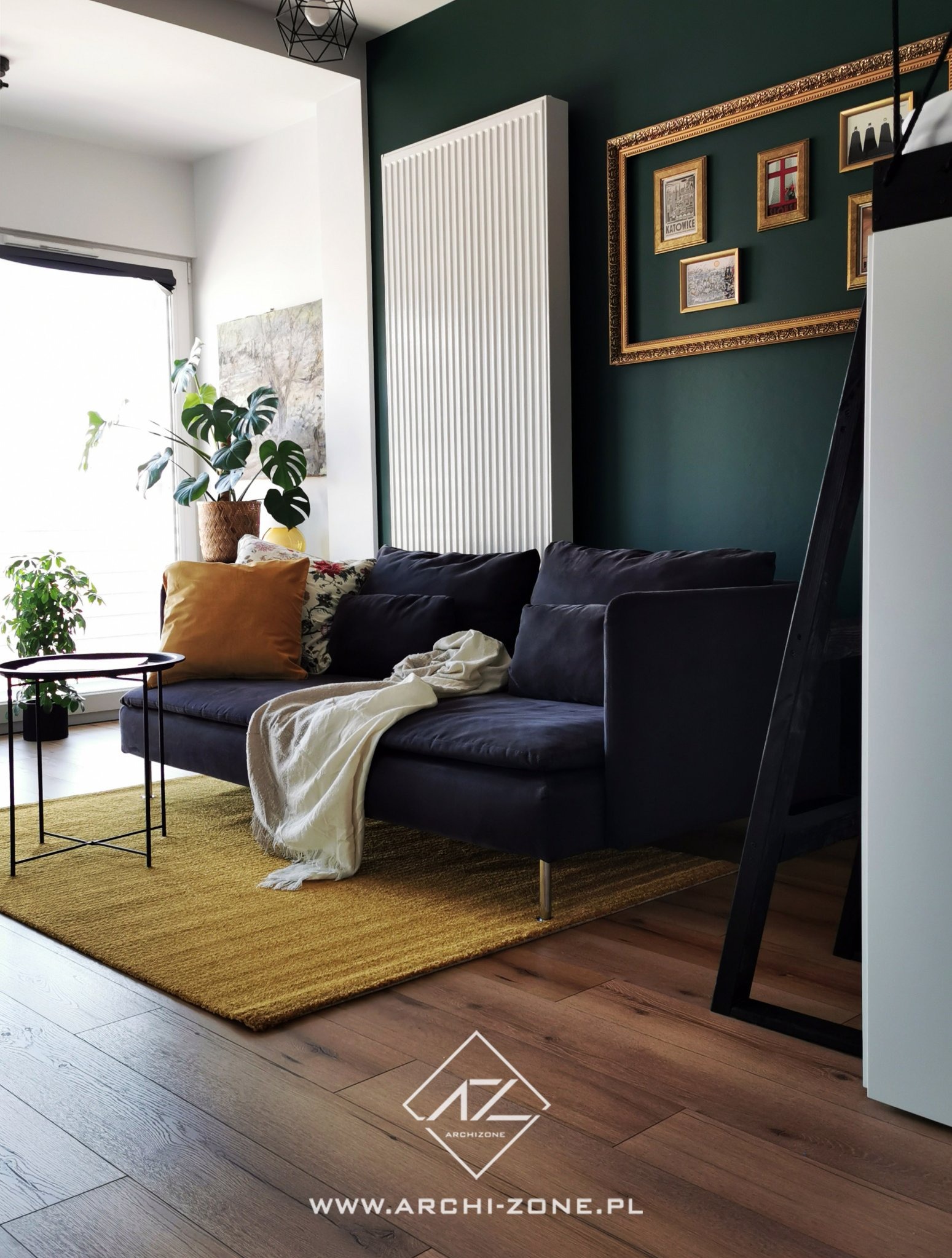 Stylowy salon z ciemną sofą, żółtym dywanem i zieloną ścianą z galerią obrazków w złotych ramach. Rośliny doniczkowe dodają świeżości wnętrzu.