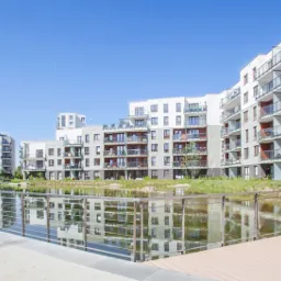 Nowoczesne osiedle apartamentowców z balkonami, odbijające się w stawie z roślinnością, widziane z drewnianego pomostu w słoneczny dzień.