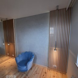 P.P.H.U WISAGE Sławomir Wierzbicki - Niebieski fotel muszelka na drewnianej podłodze, na tle szarej ściany z dekoracyjnymi panelami lamelowymi i wiszącymi lampami.