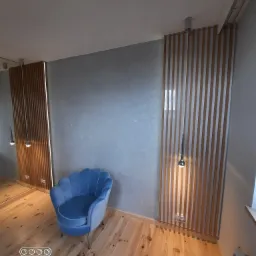 Niebieski fotel muszelka na drewnianej podłodze, na tle szarej ściany z dekoracyjnymi panelami lamelowymi i wiszącymi lampami.