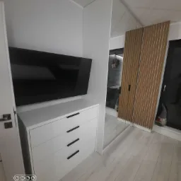 Biała szafa wnękowa z drewnianymi lamelami na drzwiach przesuwnych i komodą pod telewizorem, minimalistyczne czarne uchwyty szuflad, jasna podłoga.