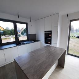 P.P.H.U WISAGE Sławomir Wierzbicki - Nowoczesna, minimalistyczna kuchnia z białymi frontami szafek, szarym blatem i wyspą kuchenną, widok z perspektywy podłogi, okno z widokiem na ogród.