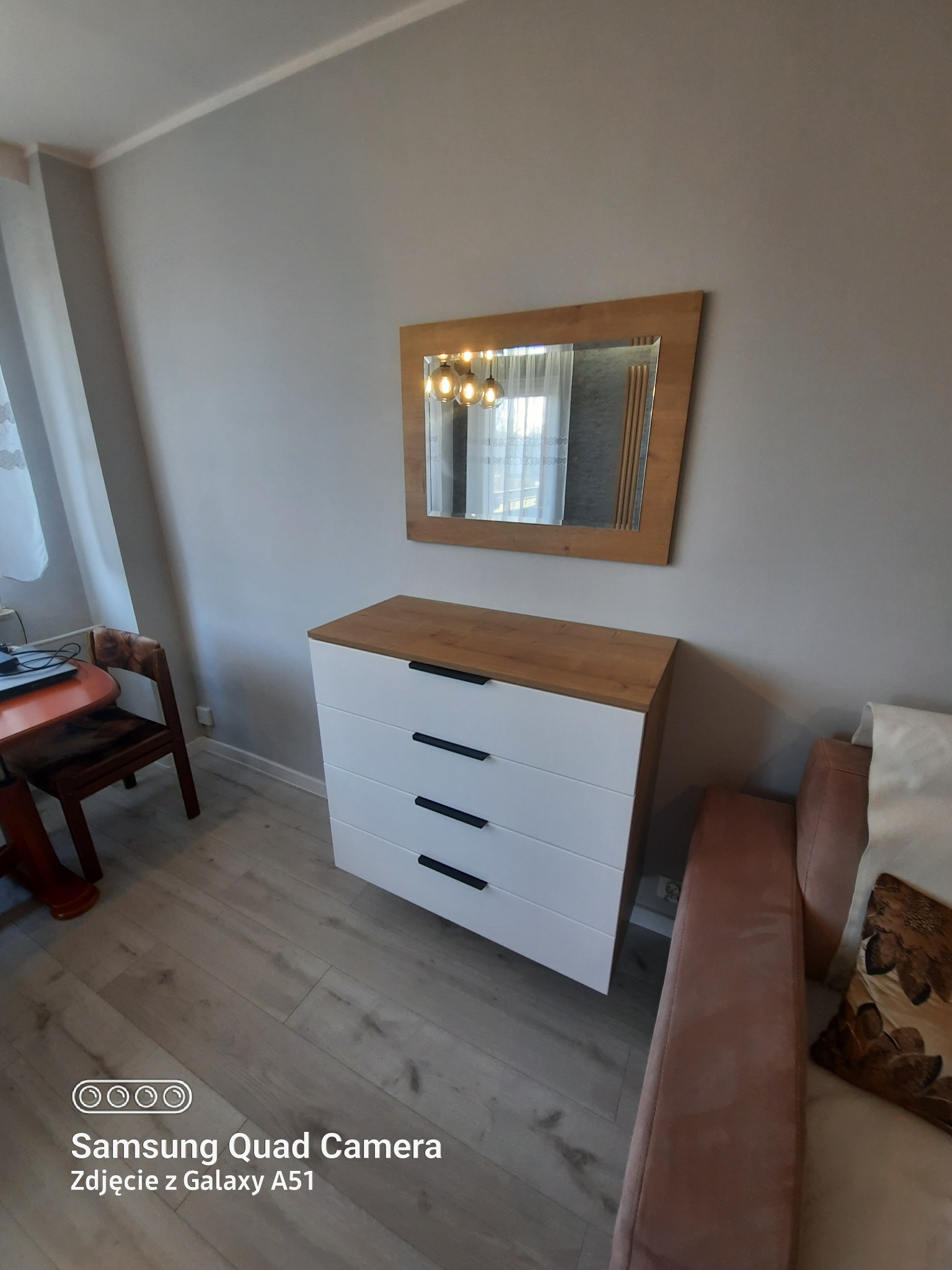 Biała komoda z czterema szufladami i drewnianym blatem, wiszące lustro w drewnianej ramie, widok na fragment pokoju z sofą i stolikiem.