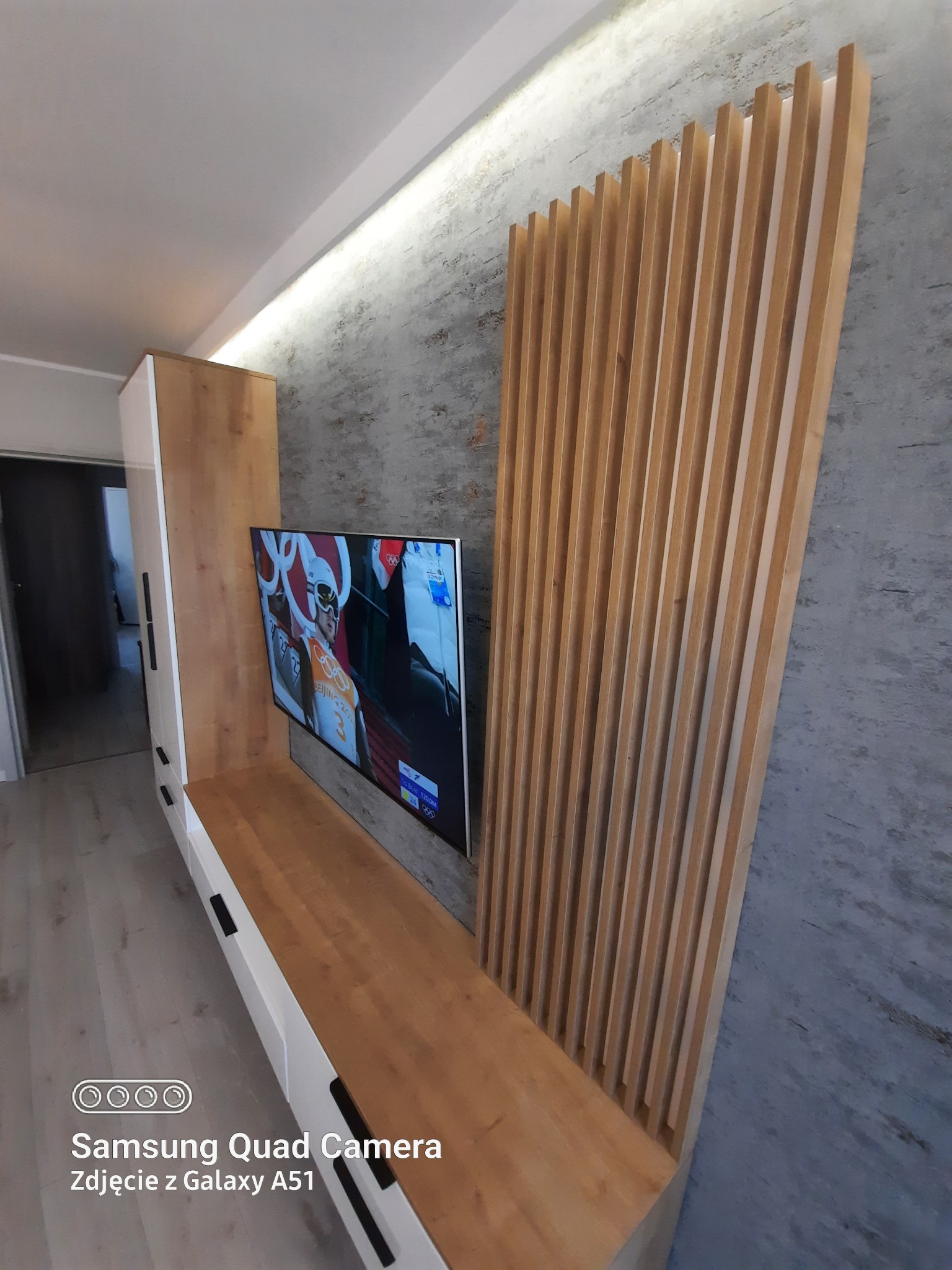 Nowoczesna zabudowa ściany z telewizorem: biało-drewniana szafka RTV z czarnymi uchwytami, drewniane lamele dekoracyjne na tle szarej ściany strukturalnej, podświetlenie LED podsufitowe.