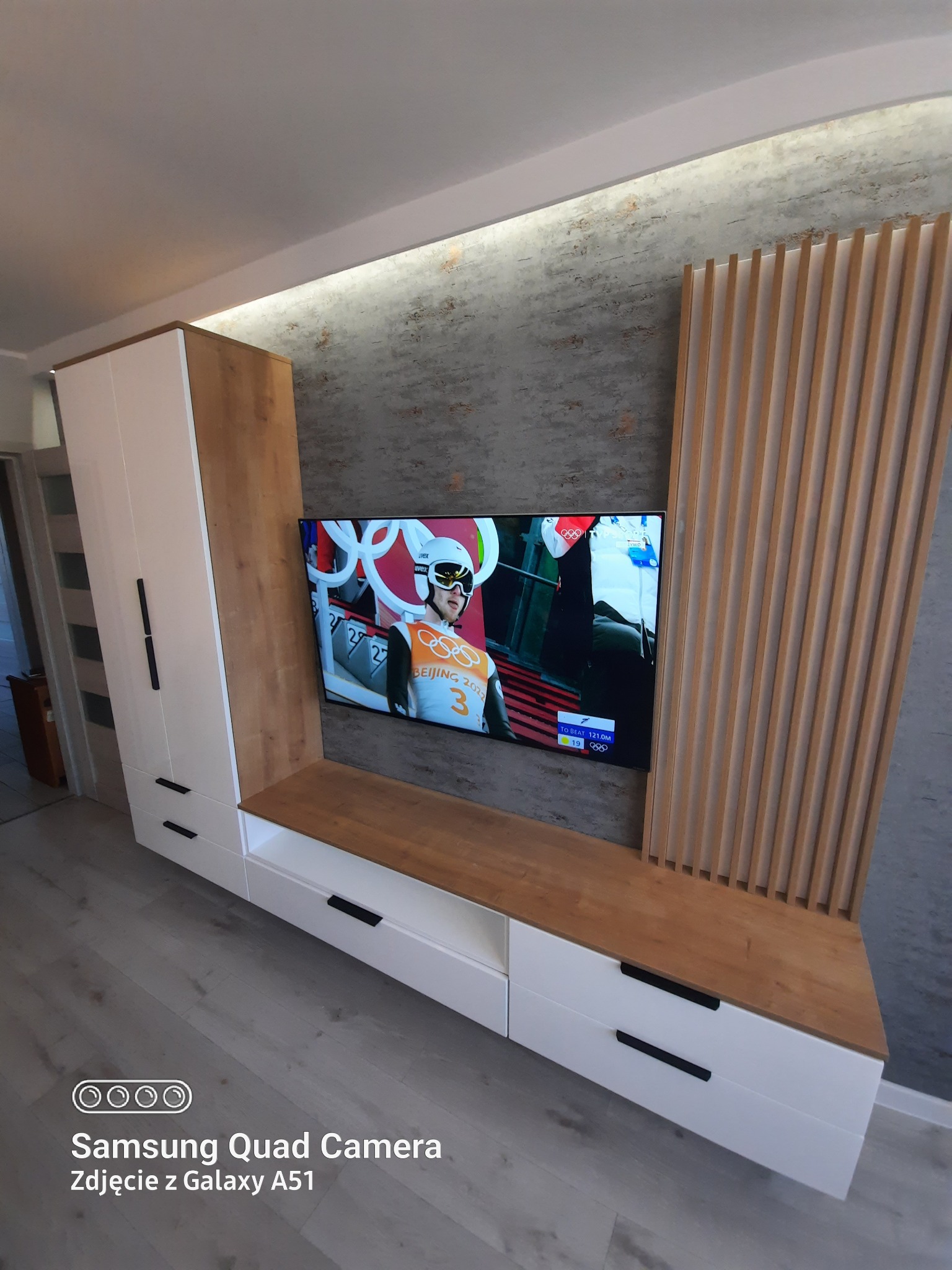 Nowoczesna szafka RTV na wymiar z białymi frontami, drewnianym blatem i pionowymi lamelami dekoracyjnymi na tle ściany imitującej beton, z telewizorem wyświetlającym zawody sportowe.