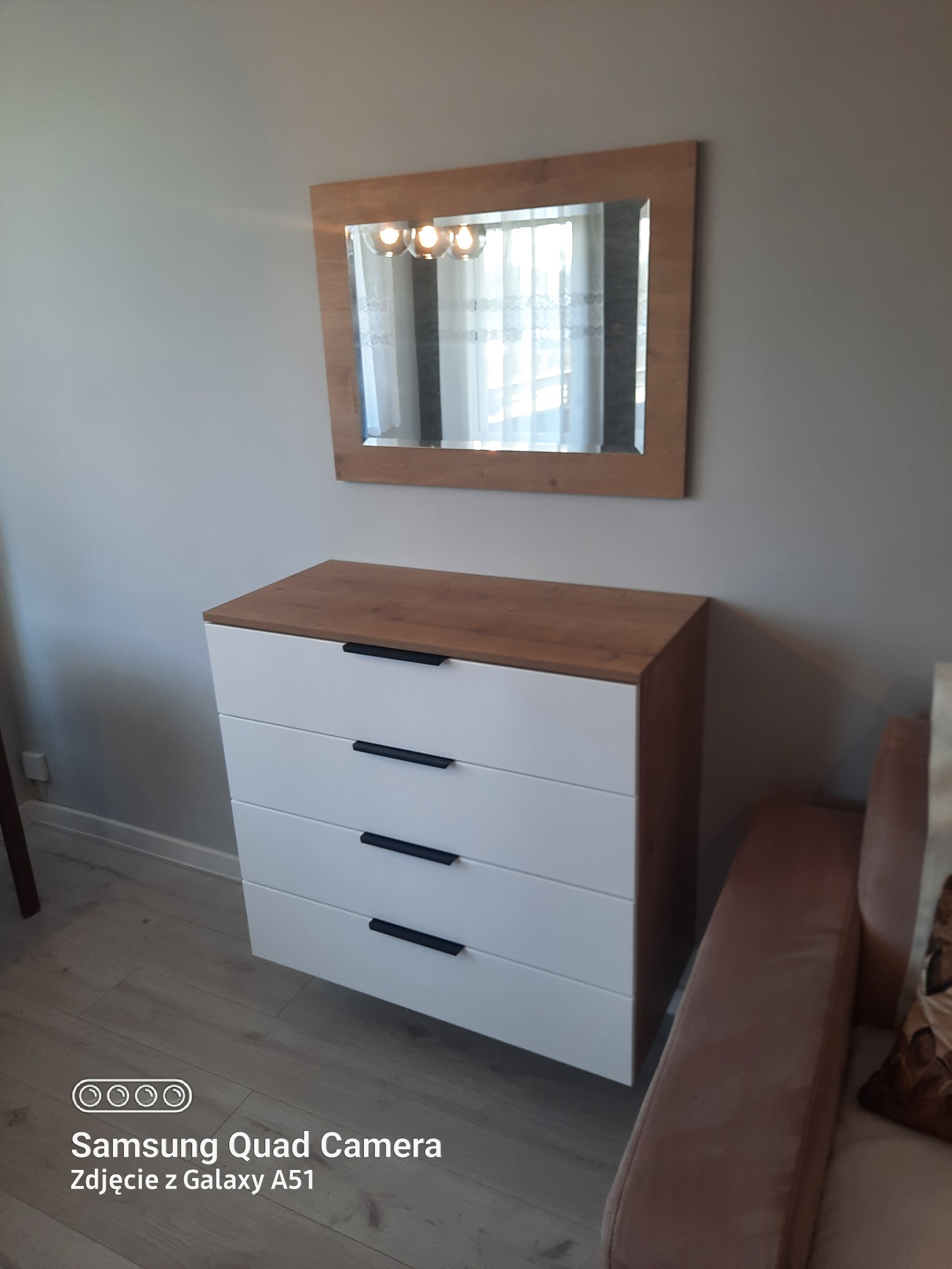 Komoda z czterema białymi szufladami i drewnianym blatem, nad nią lustro w drewnianej ramie, odbijające lampę sufitową.