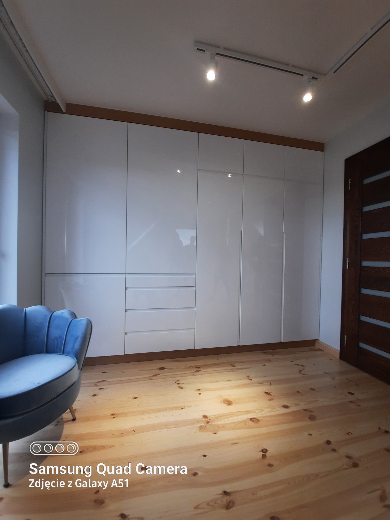 Biała szafa wnękowa z połyskującymi frontami, drewnianą ramą i wbudowanymi szufladami, oświetlona reflektorami na suficie, z drewnianą podłogą i błękitnym fotelem w stylu retro.