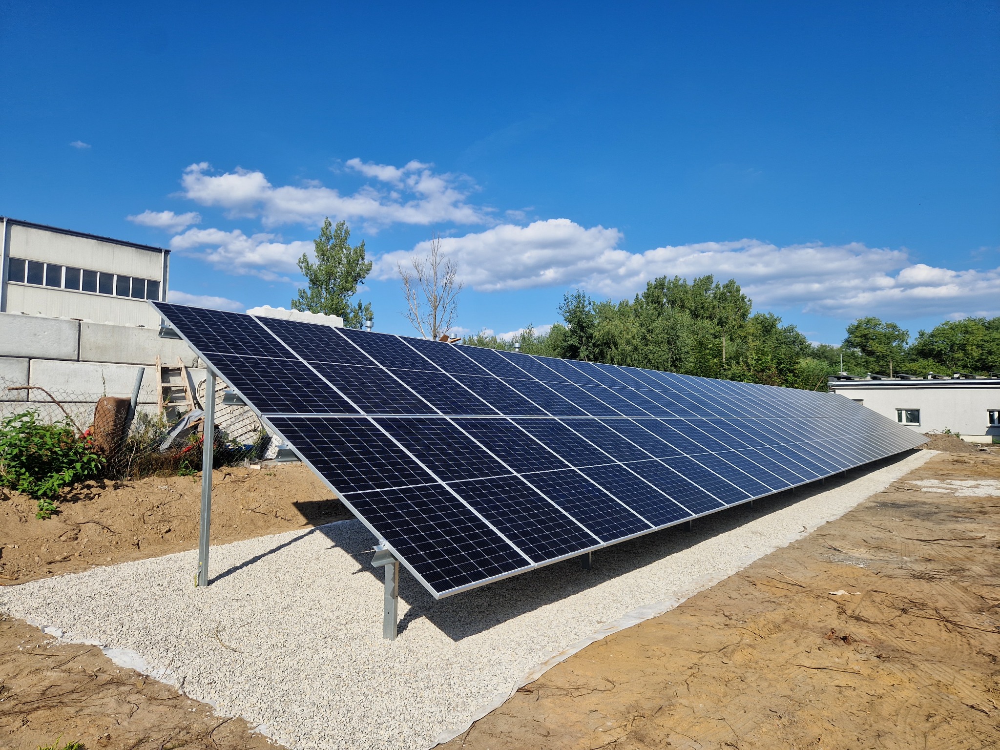 Rząd paneli fotowoltaicznych na metalowej konstrukcji, zamontowany na żwirowym podłożu, z budynkiem w tle pod błękitnym niebem.