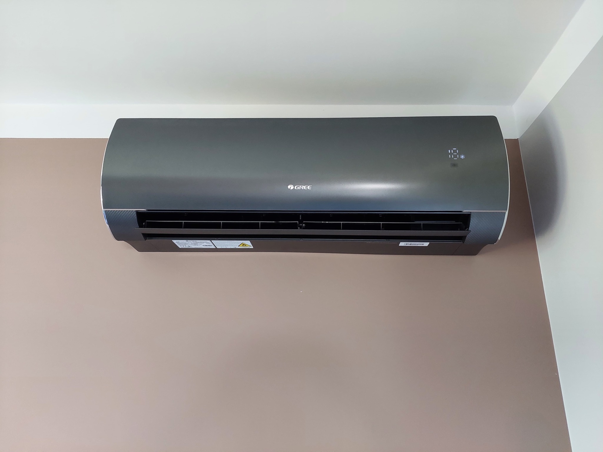 Szary klimatyzator marki Gree zamontowany na ścianie w pomieszczeniu, widoczny wyświetlacz temperatury i logo producenta.